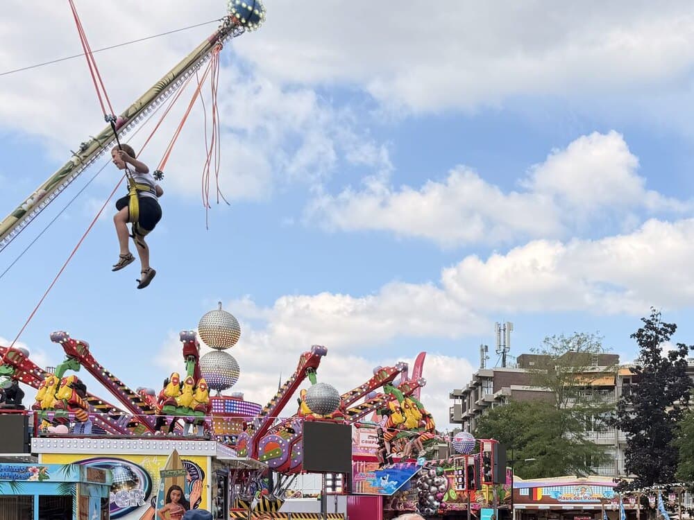 Een zweefmolen op een kermis tegen een blauwe lucht met wolken, met daaronder de kleurrijke opbouw van de attracties.